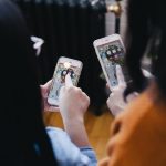 Two ladies viewed from behind point fingers at their mobile phones displaying apps