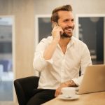 Man in white shirt talks on mobile phone seated in front of laptop computer on a desk