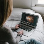 Woman sits on edge of bed working on laptop