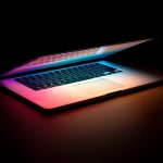 Half open laptop glows pink and purple hues from screen against black background