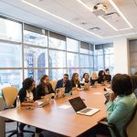 Stakeholders sit around boardroom table in boardroom with laptops open