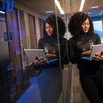 Young woman stands in office passageway leaning against glass panel while holding a laptop in her hands