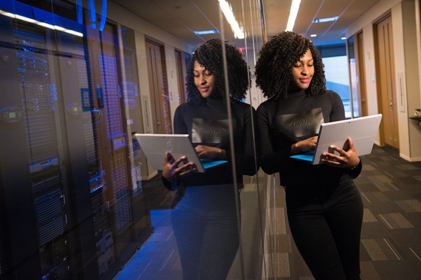 Young woman stands in office passageway leaning against glass panel while holding a laptop in her hands