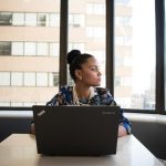 Young woman sits in front of laptop in office looking out of the window at city skyscrapers