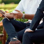Two businessmen sit on a bench outside discussing something on a tablet screen
