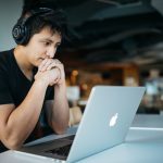 Man sits in front of laptop screen with headphones on his head inside an office with a view of a nightscape