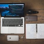 Open laptop seen from above rests on a rough wooden desk next to a diary two smartphone keys nails and a camera